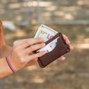A person's hands holding an open dark brown leather wallet with cash and cards visible, photographed outdoors against a blurred natural background.