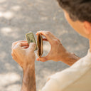 A close-up lifestyle shot of a man holding a folded dollar bill and a brown leather wallet, viewed from behind as he looks down at the items against a blurred outdoor background.